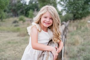 Young girl in white dress poses outdoors in a natural setting with grass and trees in the background.