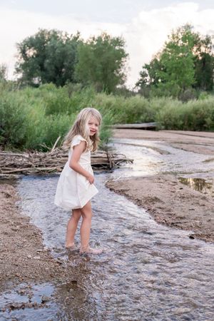 A small child in a white dress splashes and plays in a shallow stream on a summer day near trees and greenery.