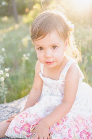 Young child in white dress sitting in sunlit meadow with delicate pink floral details.