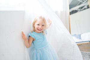 Child in light blue tulle dress playing with white curtains in bright room.