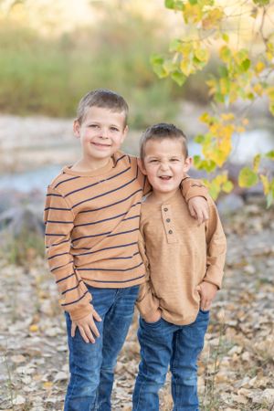 Two children in casual sweaters smiling together outdoors with autumn leaves in the background.