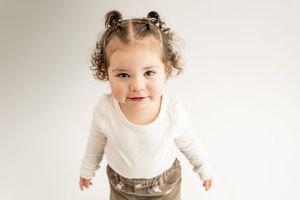 A toddler on a white background wearing a flowy off-white dress smiles brightly at the camera.