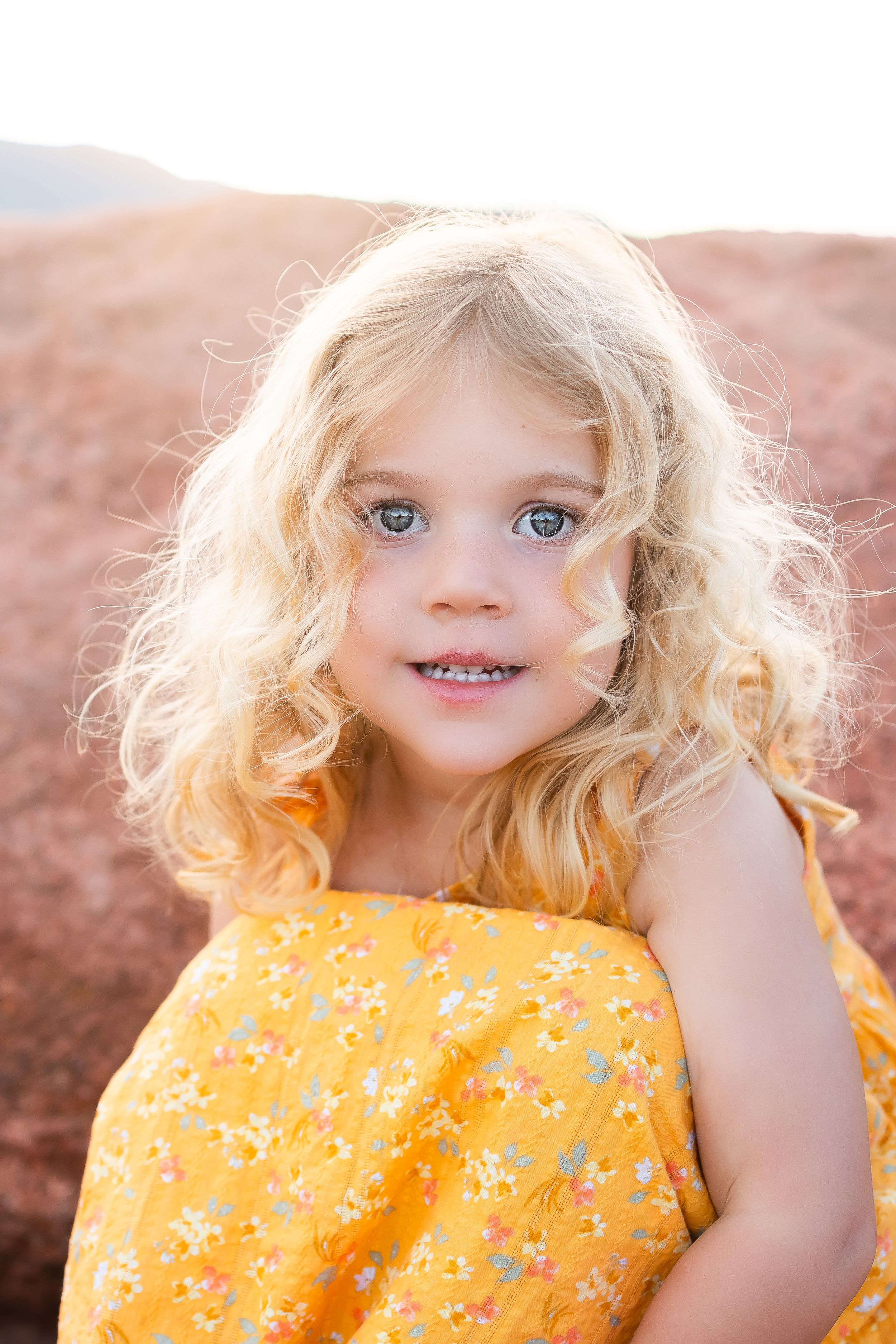 Young child in yellow floral dress with blonde curls and blue eyes smiling outdoors in warm sunlight.
