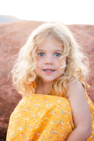 Young child in yellow floral dress with blonde curls and blue eyes smiling outdoors in warm sunlight.