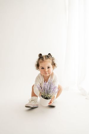 A child in a white dress lies on the floor with purple lavender flowers in a bright, airy studio setting.