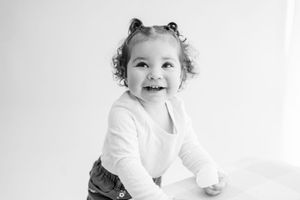 A joyful toddler in a white sweater sits on a white background in this black and white portrait.