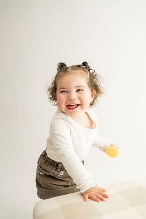 A happy child wearing a white long sleeve shirt and brown overalls sits on a light-colored surface in a studio setting.