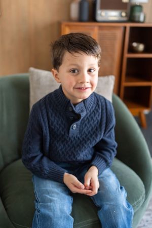 Child sitting on green armchair wearing navy cable knit sweater and jeans in cozy living room setting.