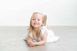 Child in white dress lying on grey floor with cheerful expression.