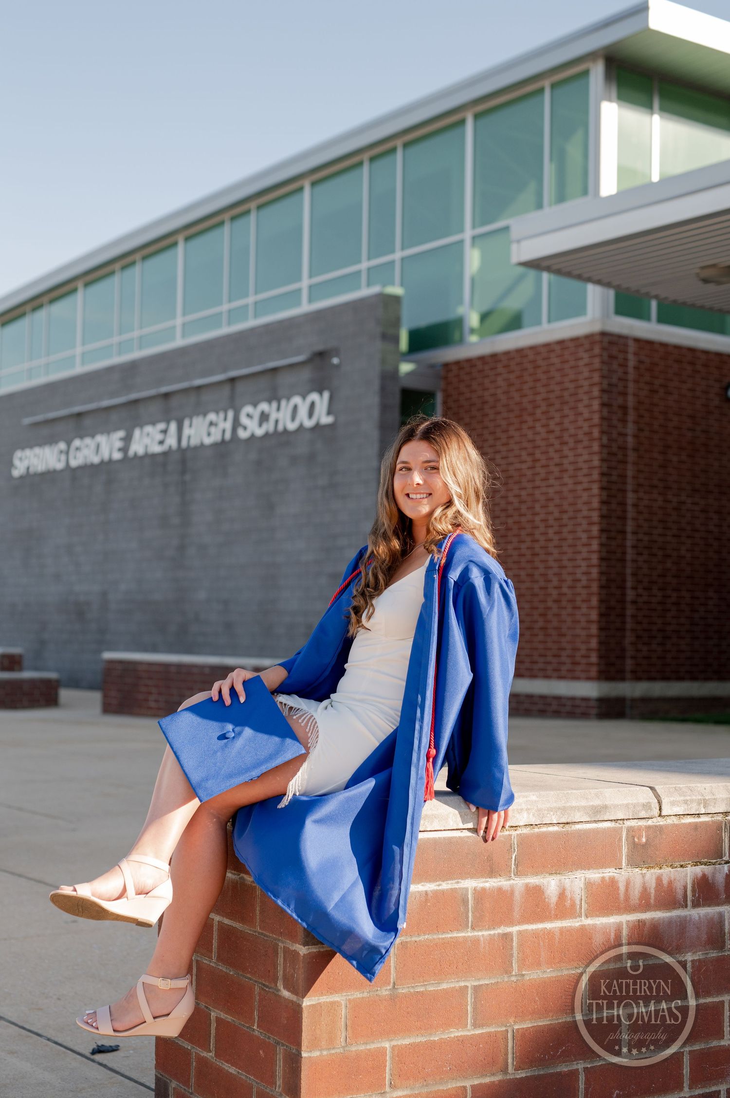 Graduate in blue cap and gown sitting on brick ledge at Spring Grove Area High School.