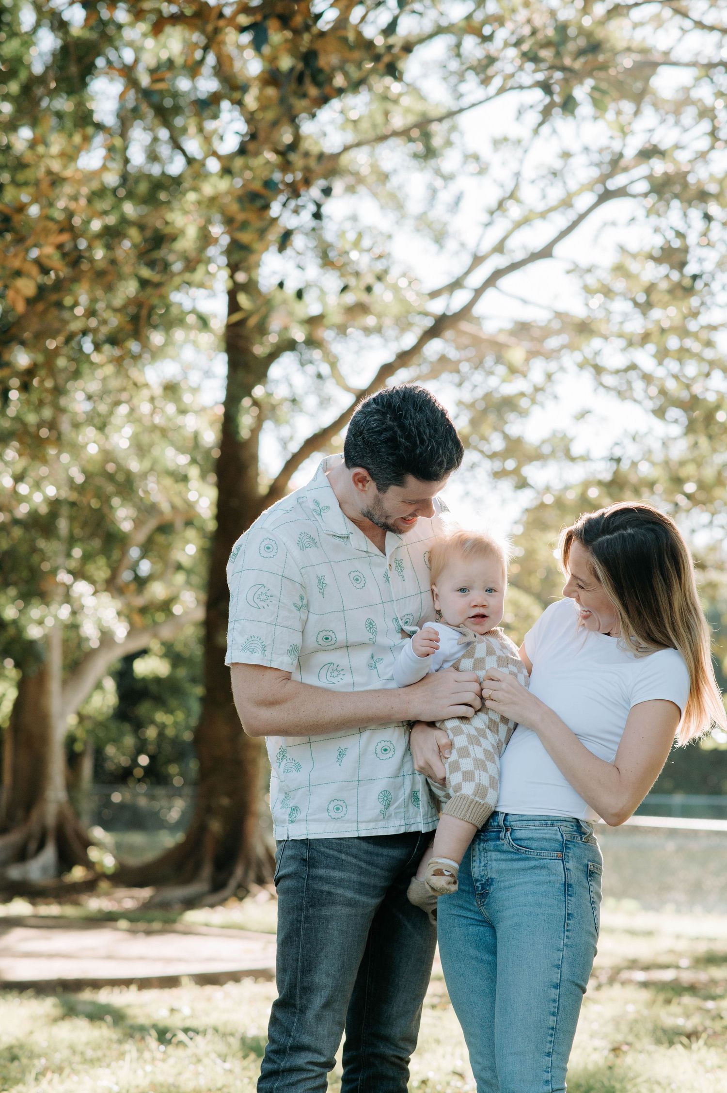 Family shares tender moment with their baby in a sun-dappled park setting.