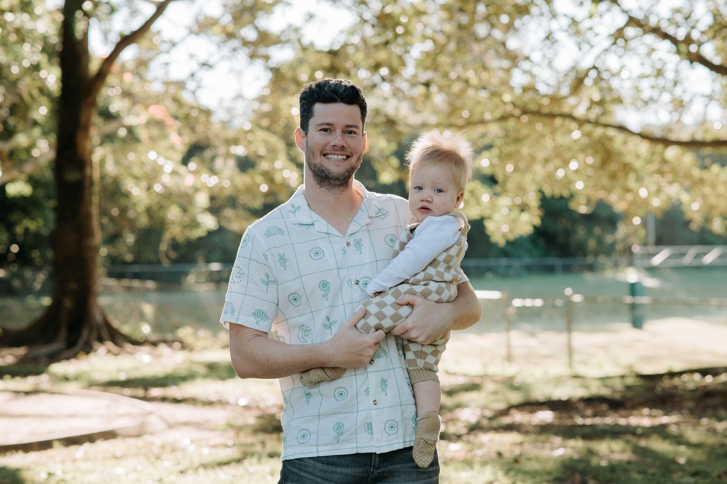 Adult holding smiling baby in patterned shirt outdoors with trees in background.