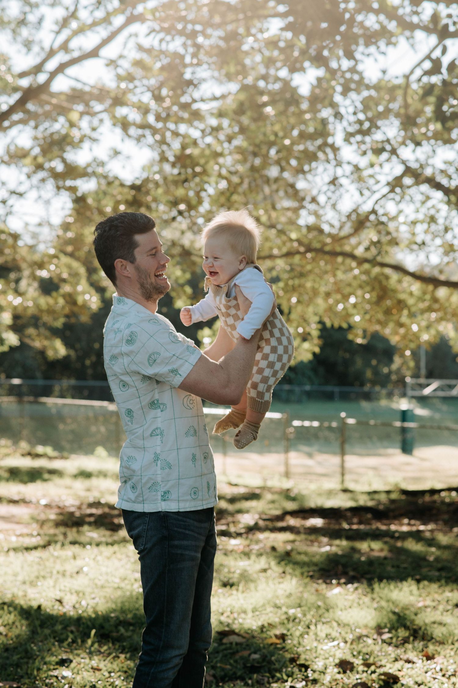 Adult lifting baby in air during sunny outdoor photoshoot near trees.