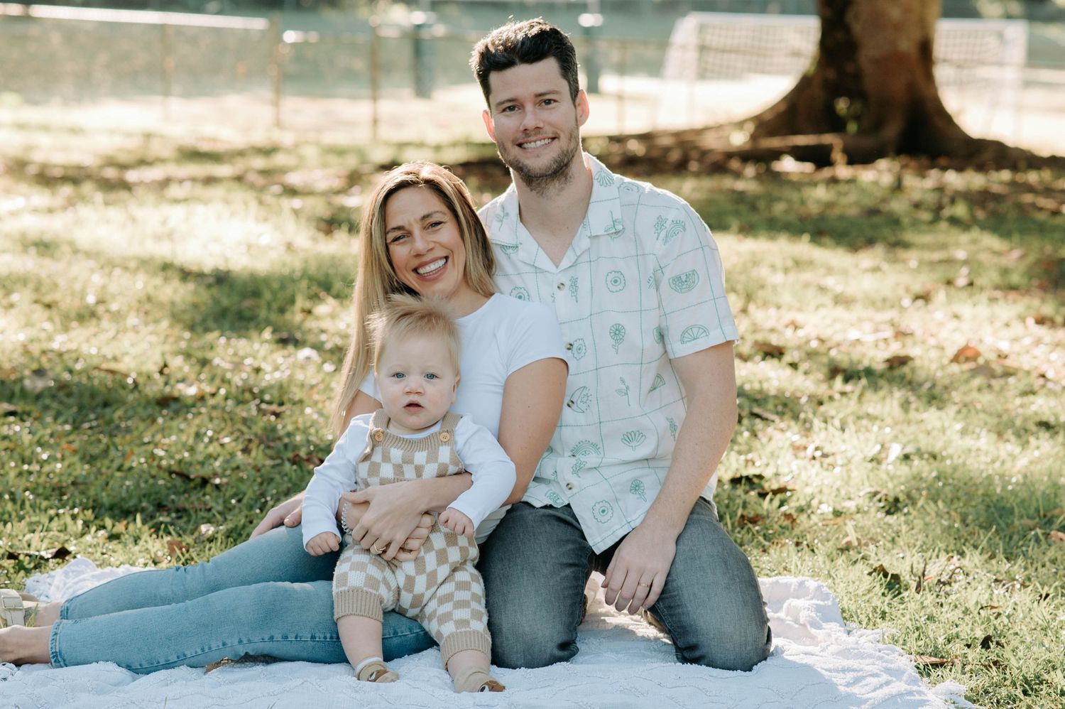 A family sits together on a blanket in a sunny park setting during a casual outdoor photo session.