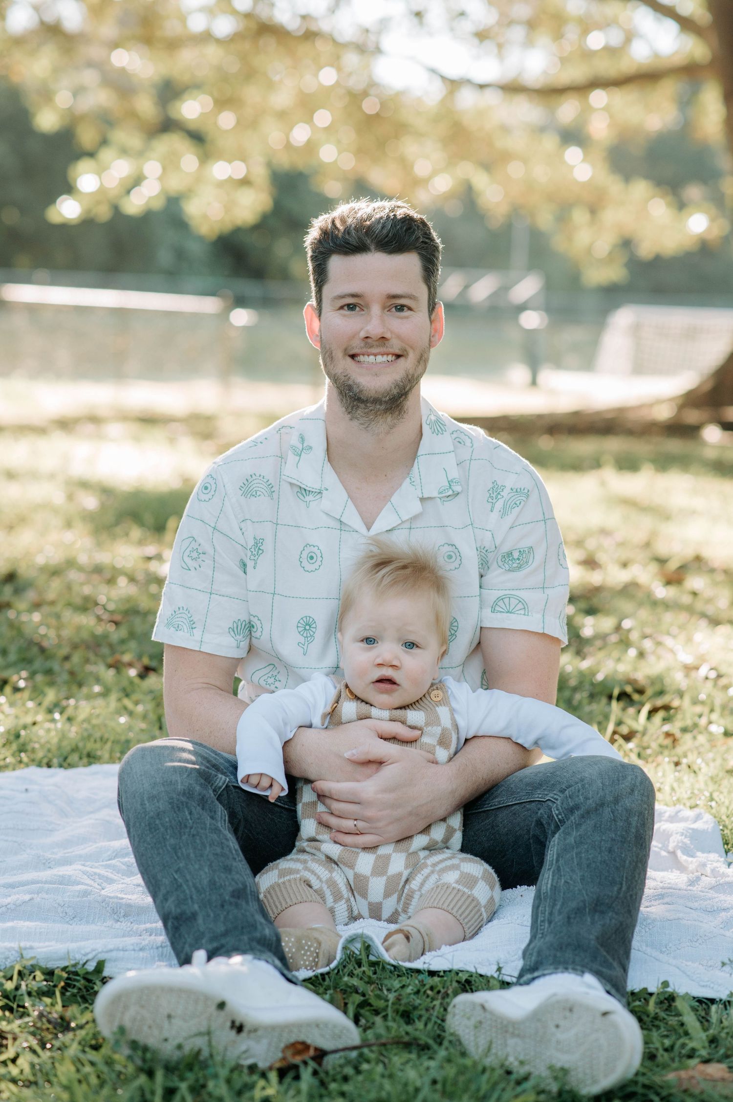 Parent and baby sitting on blanket outdoors in dappled sunlight.