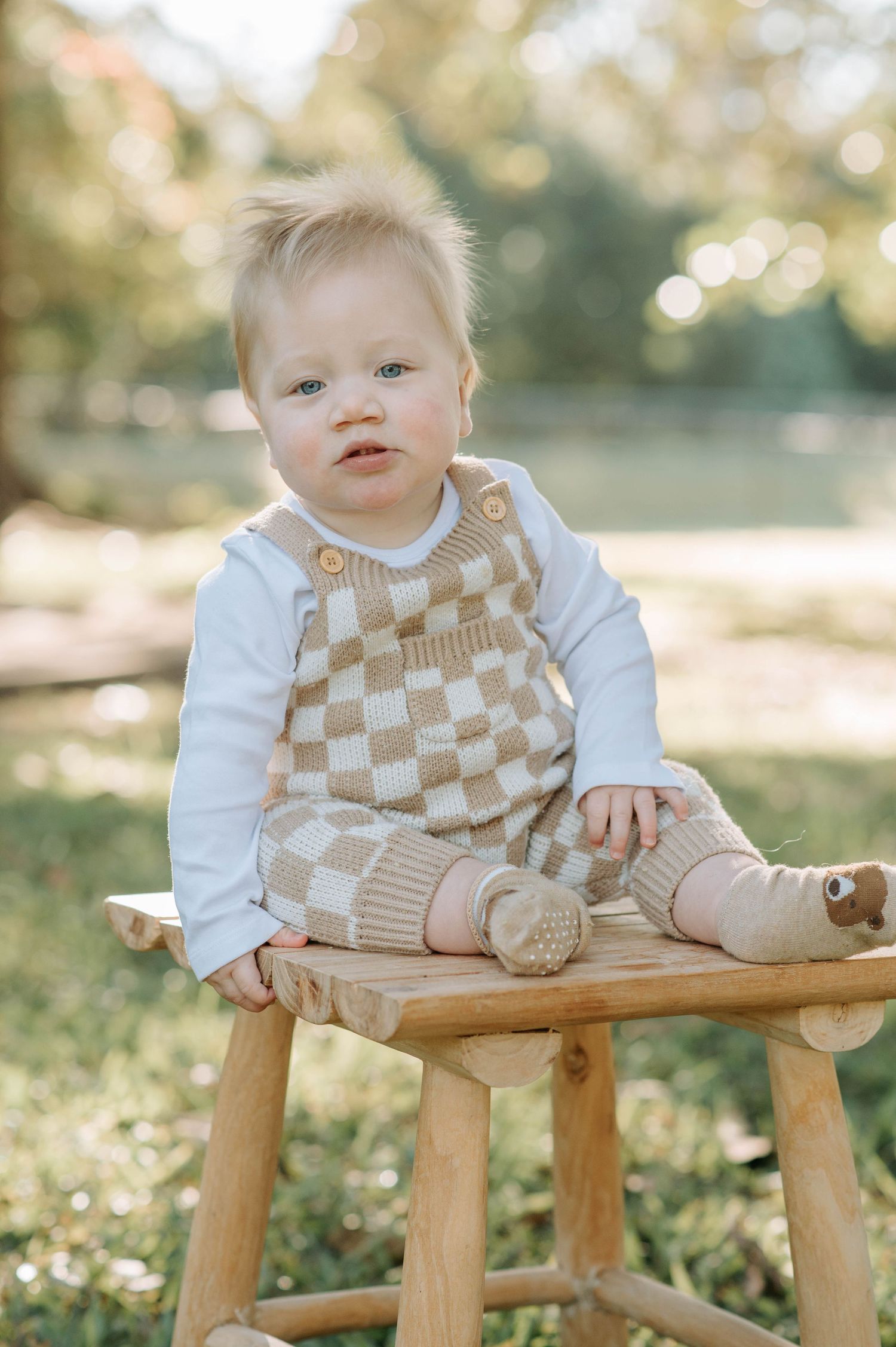 Baby wearing checkered overalls sits on wooden stool in sunlit outdoor setting.