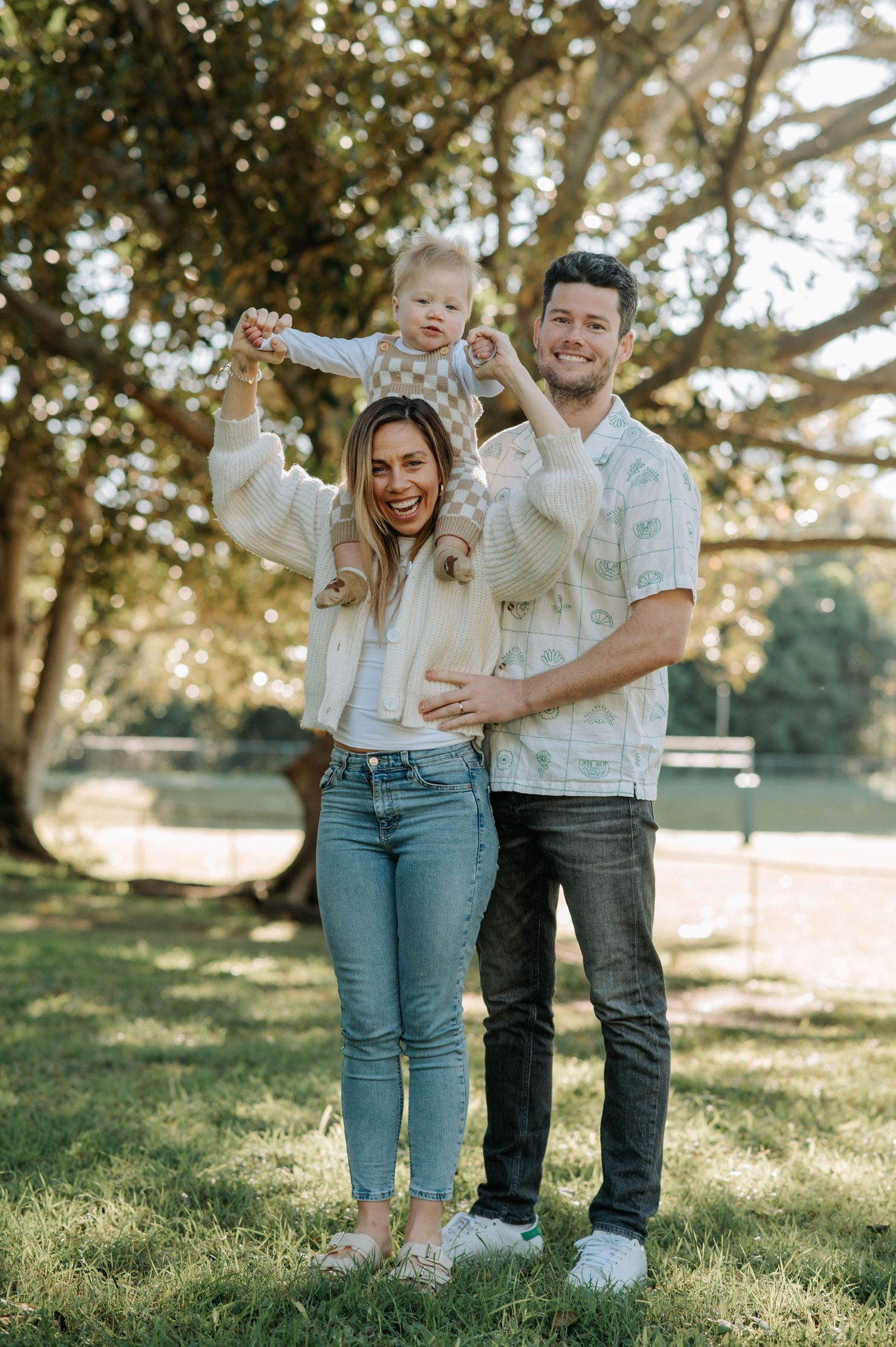 Family having fun together in park with baby lifted up in playful moment.