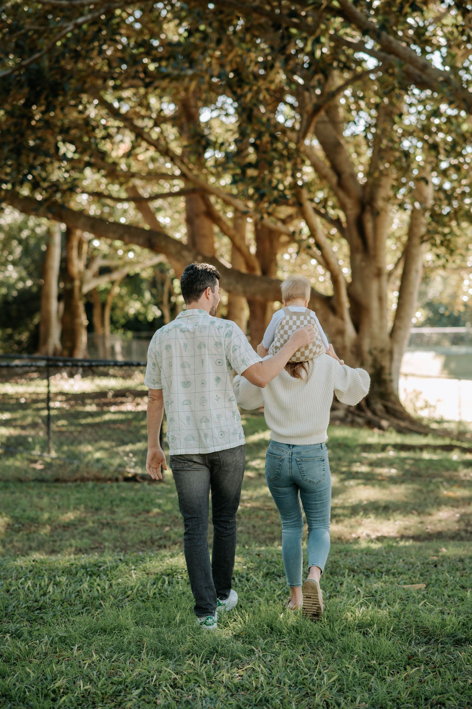 Couple walking away from camera holding hands in park under large trees.