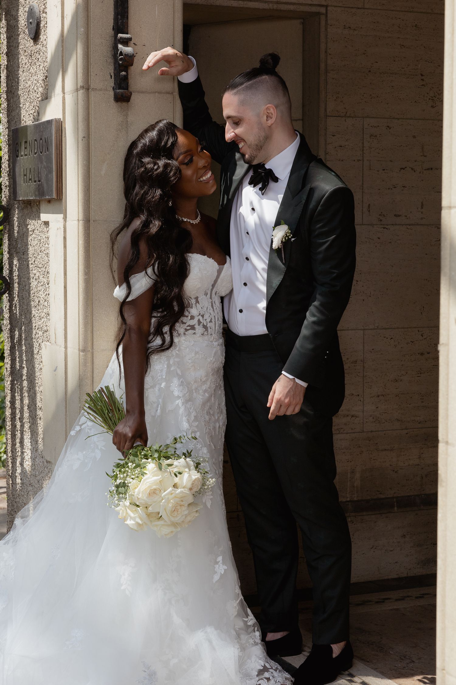 A romantic wedding photo of a couple in formal attire with the bride holding a white bouquet outside a doorway.