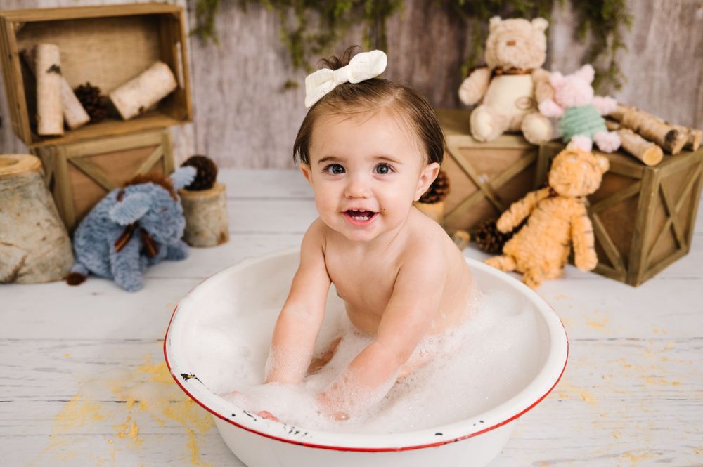 Baby sitting in a vintage enamel bathtub surrounded by stuffed animals and wooden crates.