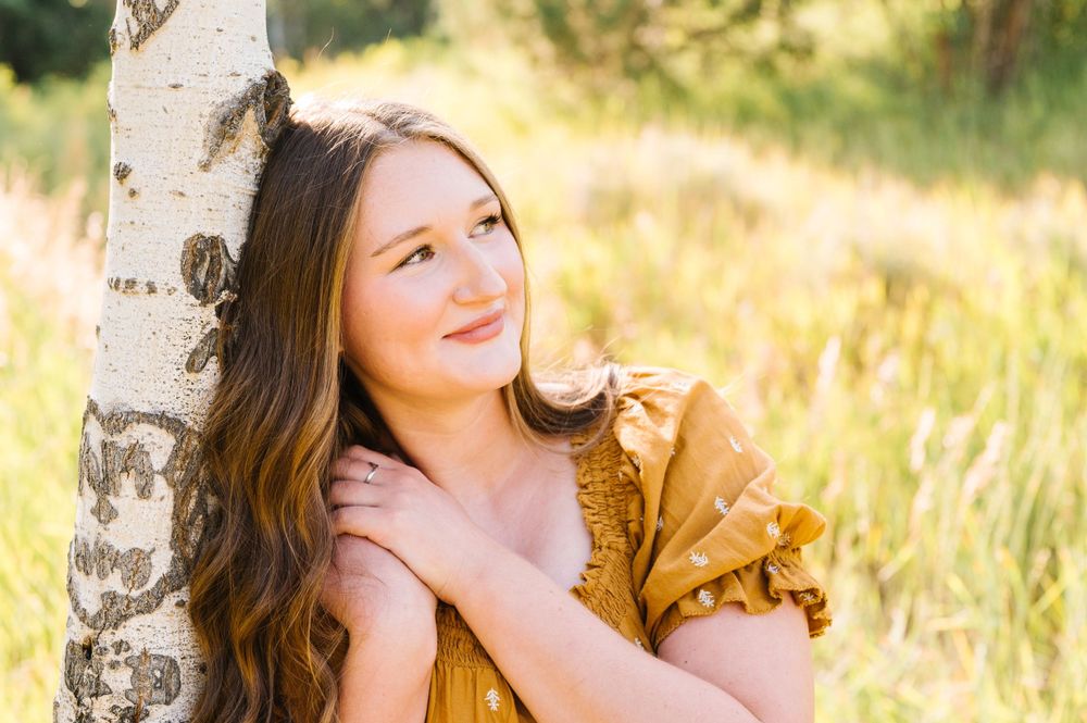 Young woman in a mustard yellow top poses thoughtfully by a birch tree in a sunlit meadow.