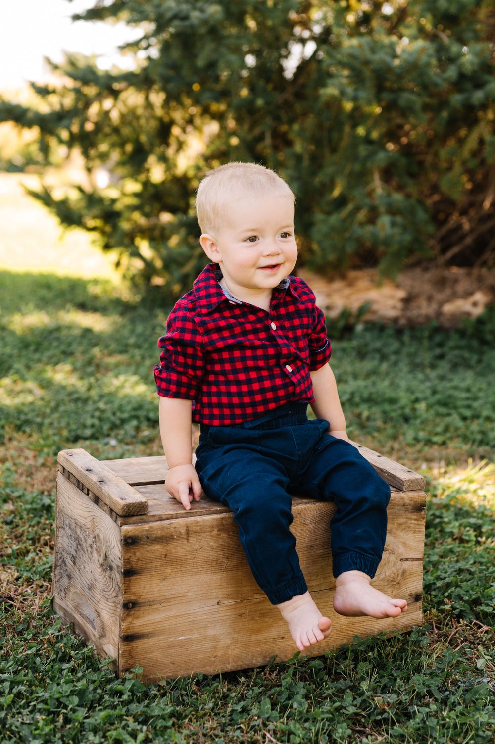 A toddler in a red plaid shirt sits on a wooden crate outdoors.