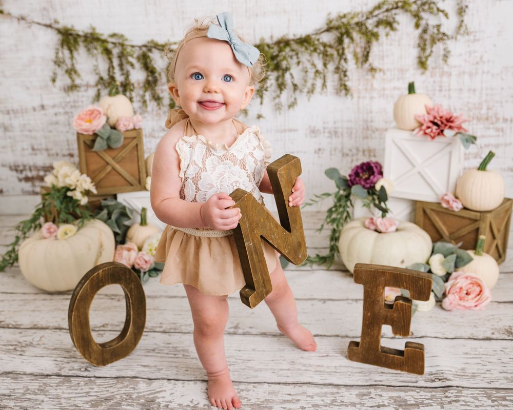 Baby in lace outfit standing near decorative 'ONE' letters with floral and vintage decor.