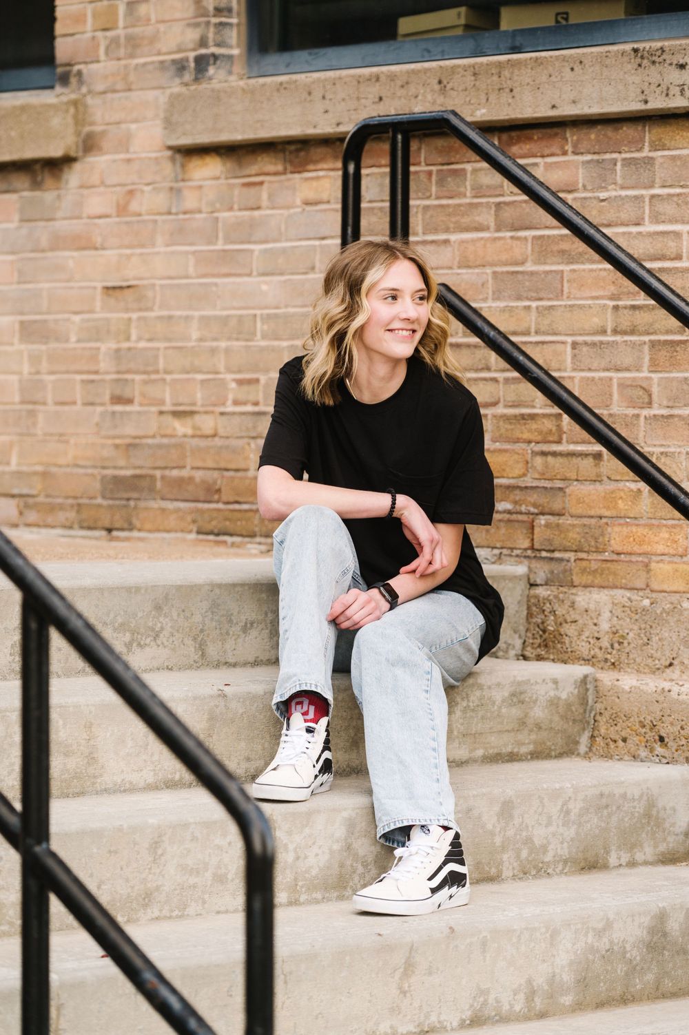 A relaxed portrait on concrete steps wearing black top, light jeans and high-top sneakers.