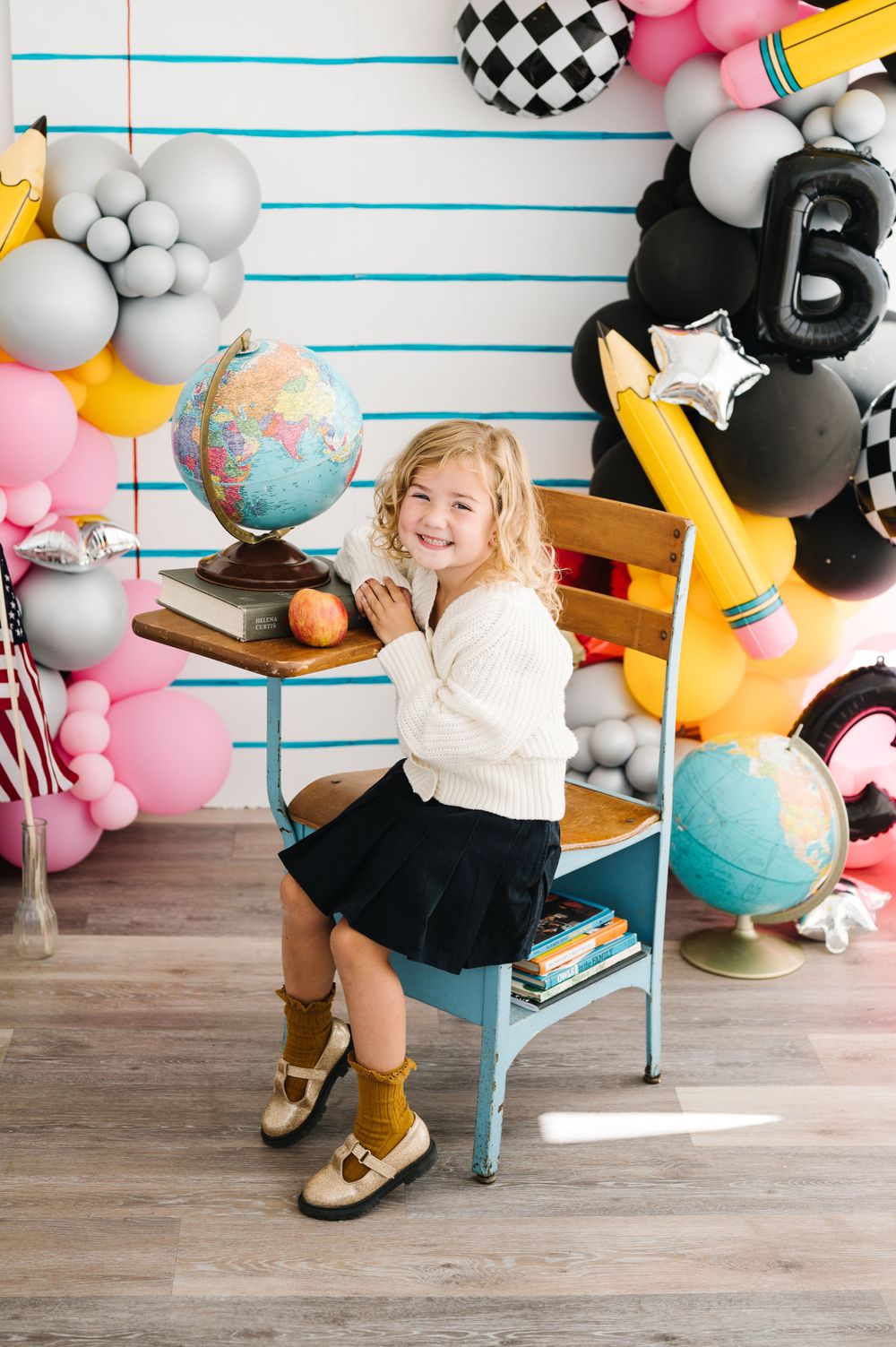 A school-themed photo setup with balloons, globes, and a young student sitting on a blue chair.