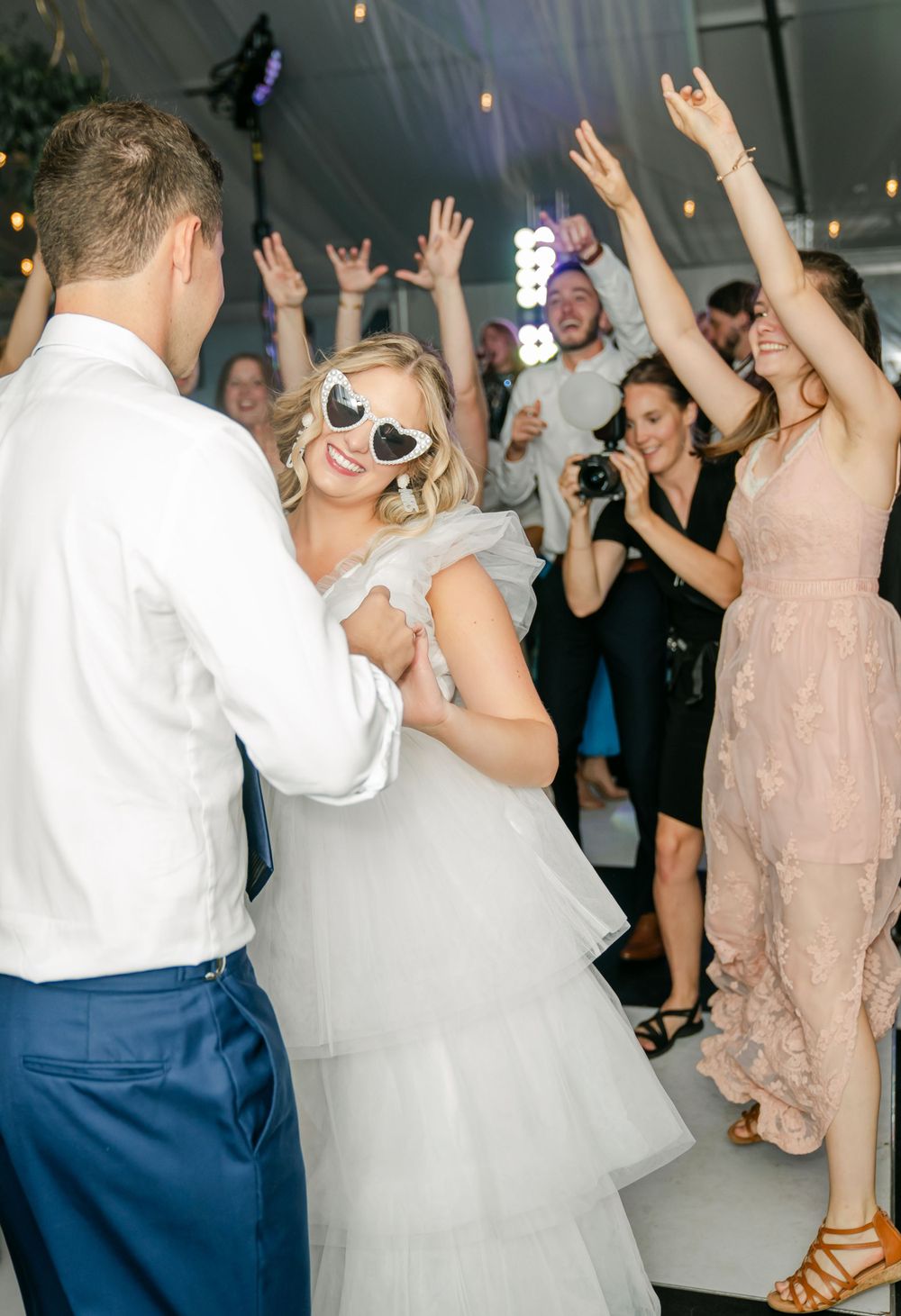 Wedding celebration on dance floor with guests dancing under string lights in a white tent.