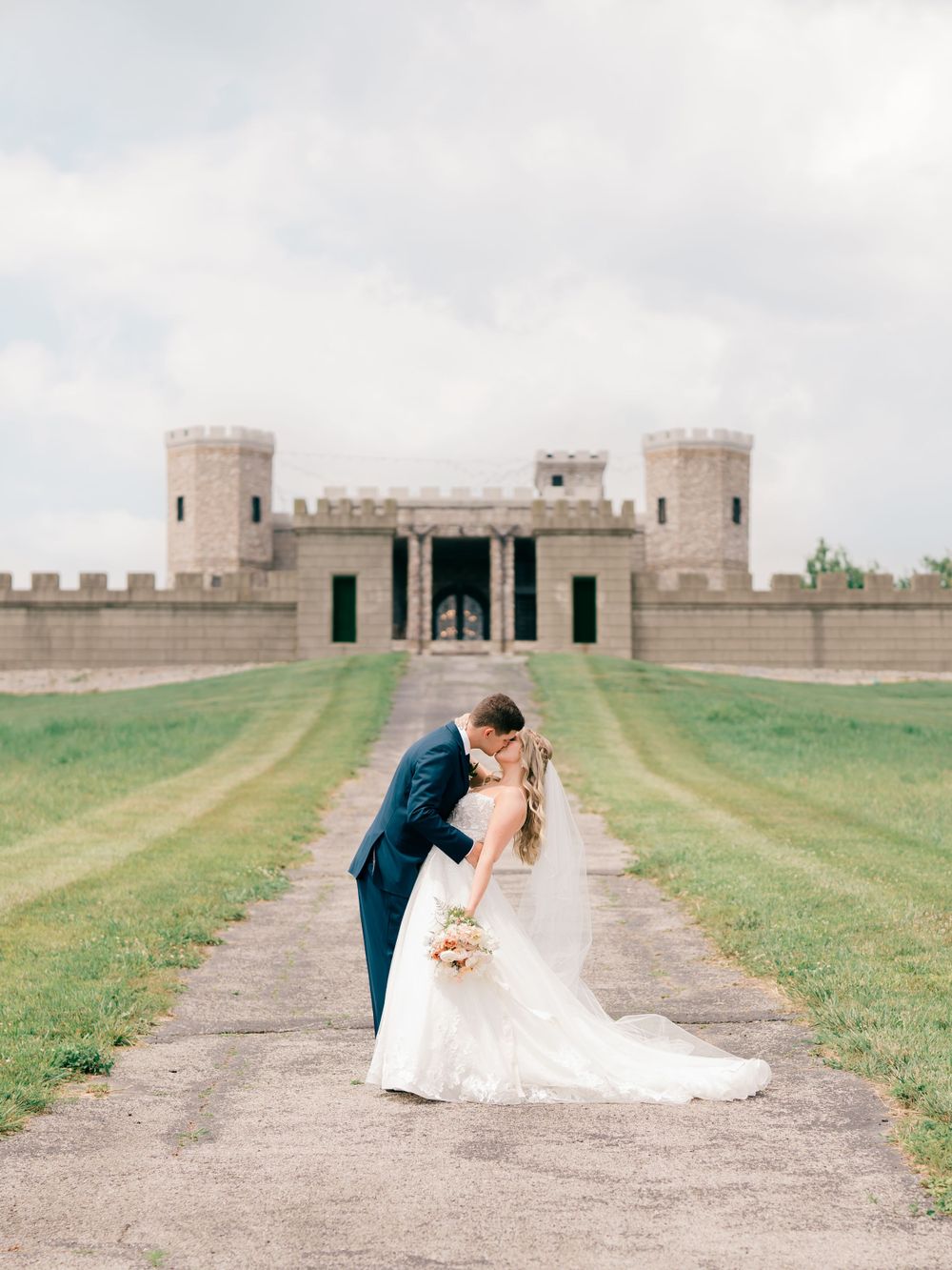 Newlyweds share romantic moment on path leading to historic castle with stone towers.