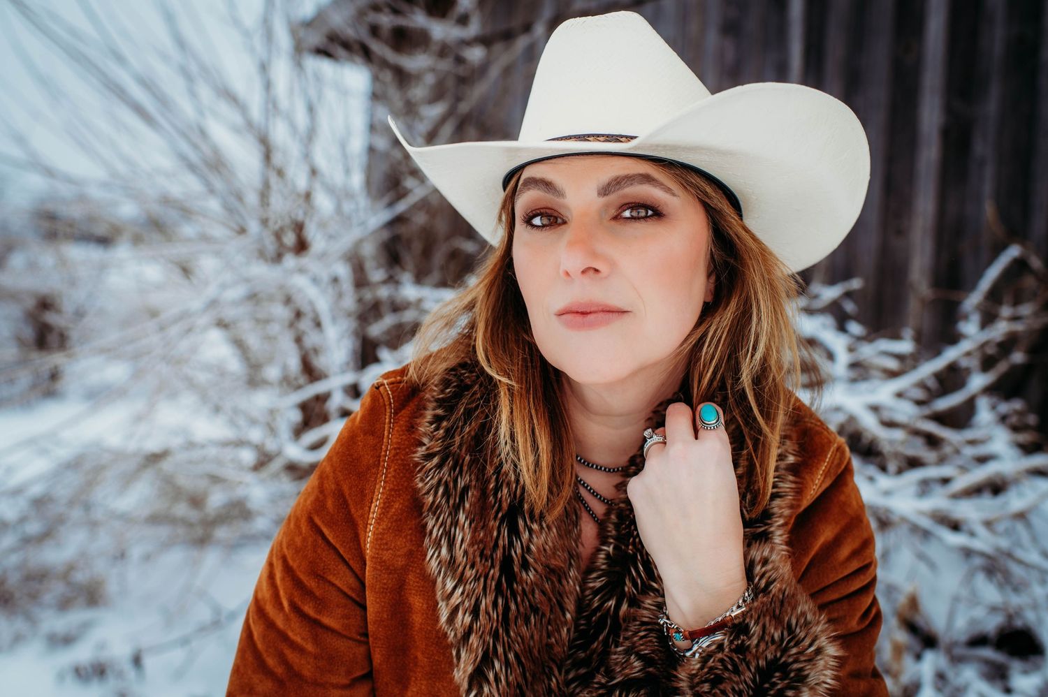 Winter portrait with model in white cowboy hat and rust-colored fur-trimmed coat against snowy background.