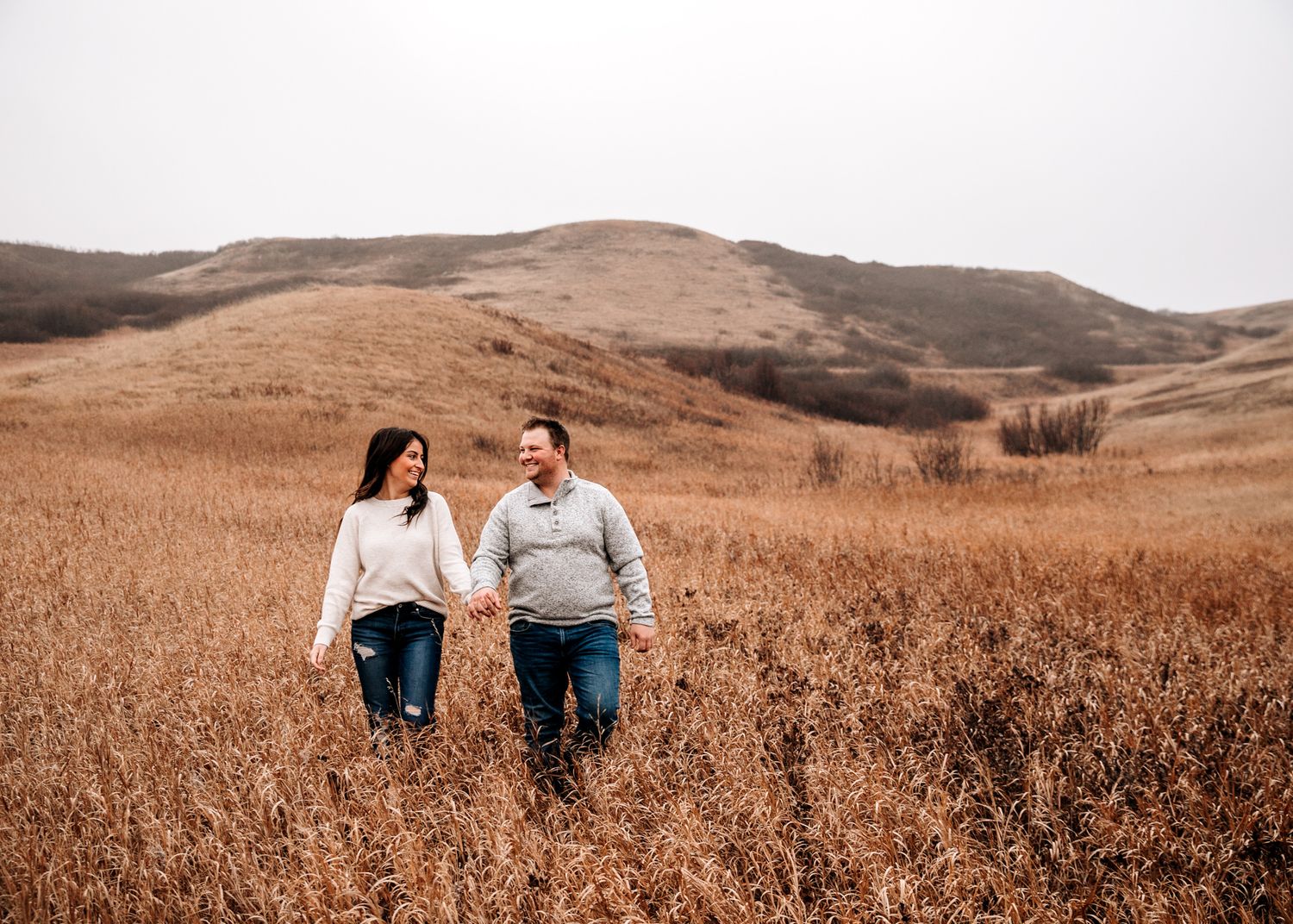 Couple holding hands and walking through a golden prairie field on an overcast day.