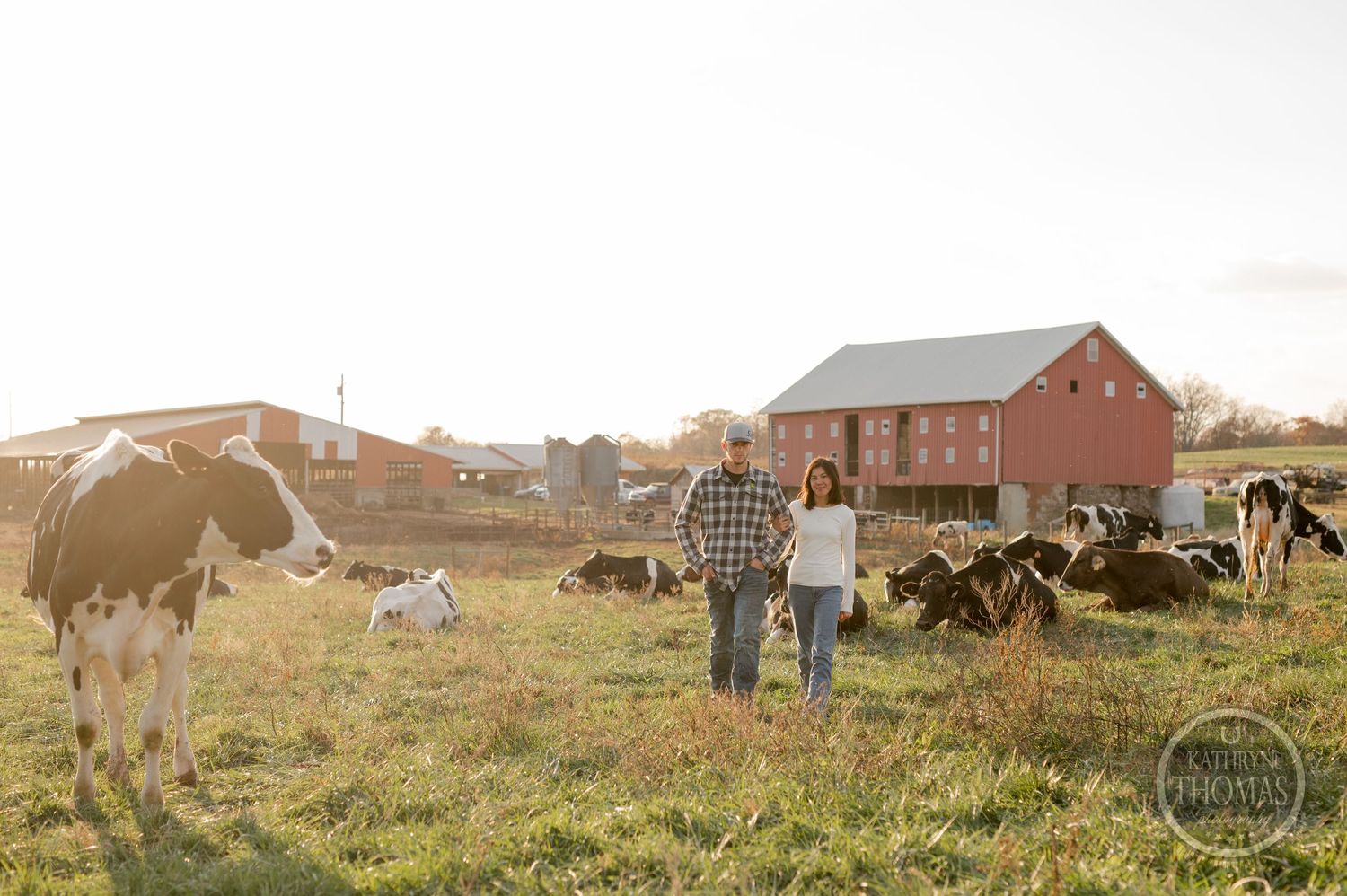 Two people stand in a pasture with dairy cows and a red barn visible in the countryside setting.