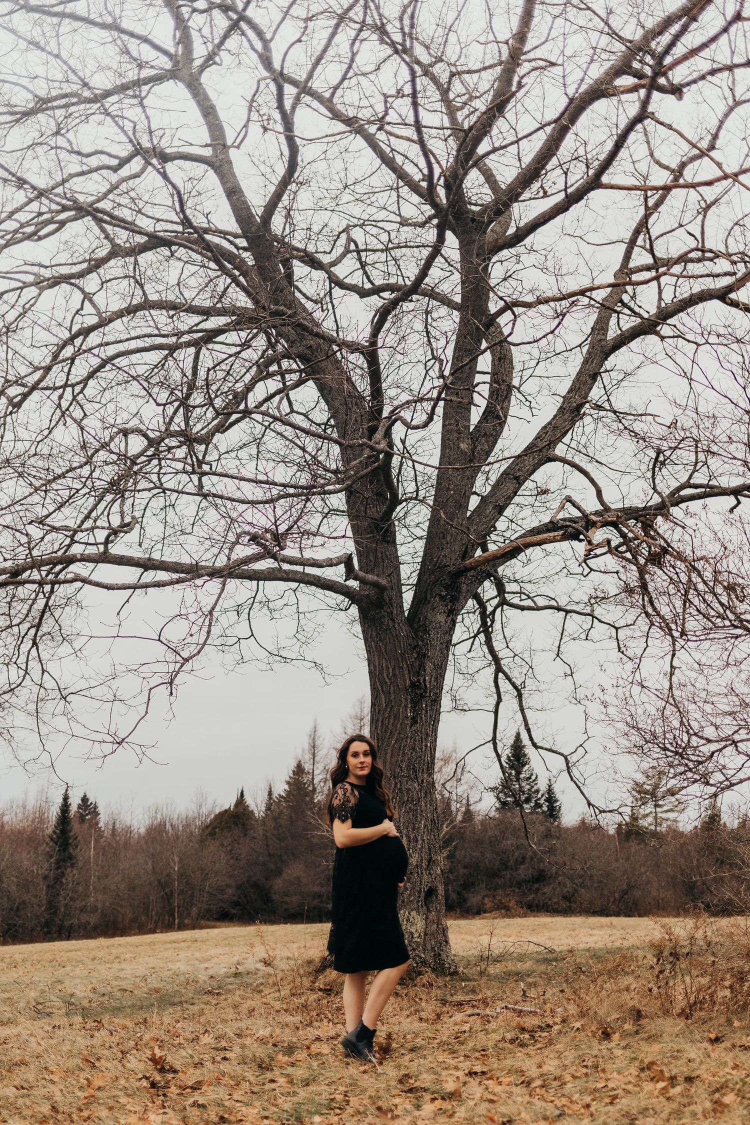 A woman in a black dress stands beside a large bare tree on a brown autumn hillside under an overcast sky.