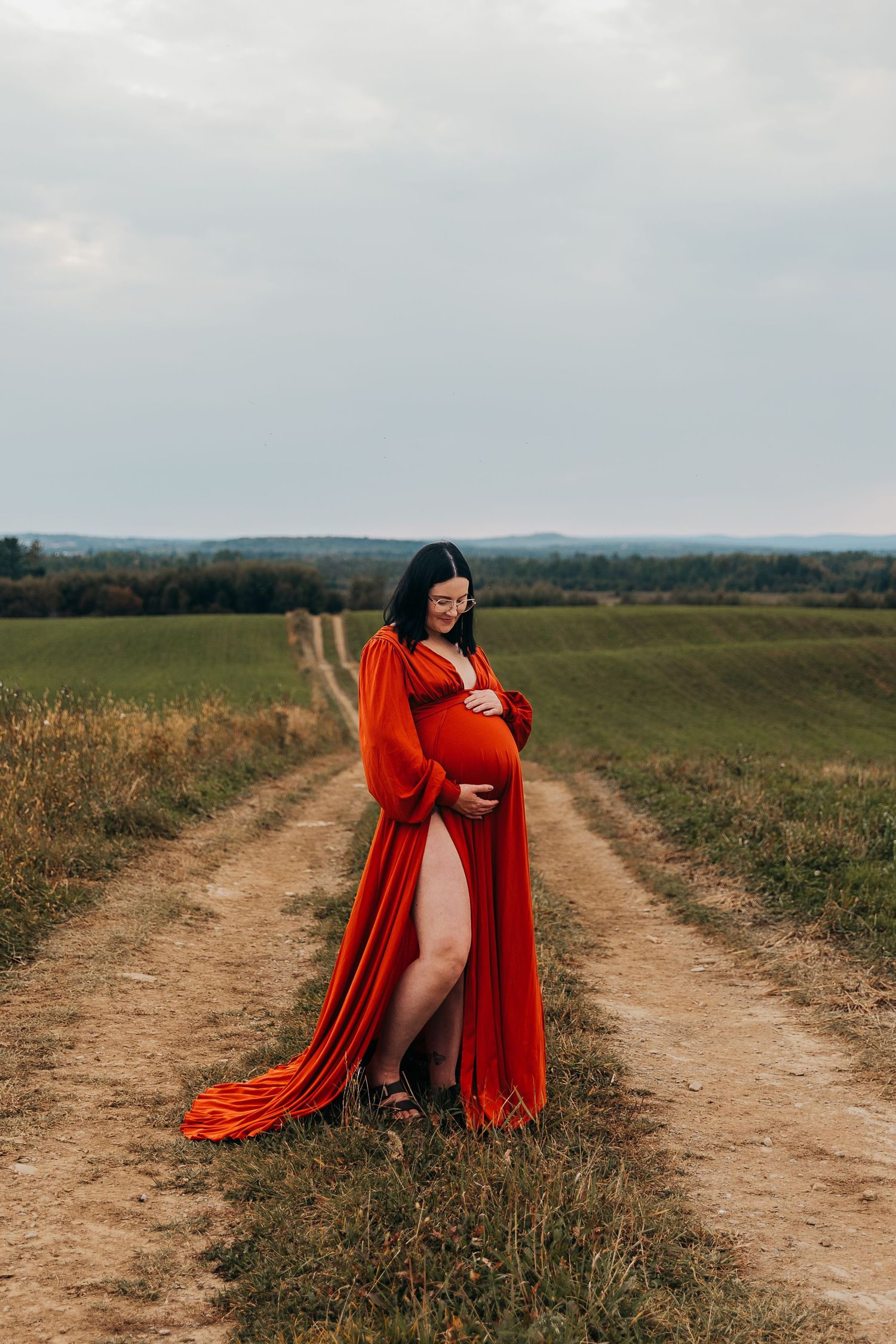 An expecting mother in a rusty orange flowing dress contrasts against a rural dirt road through fields on a cloudy afternoon.