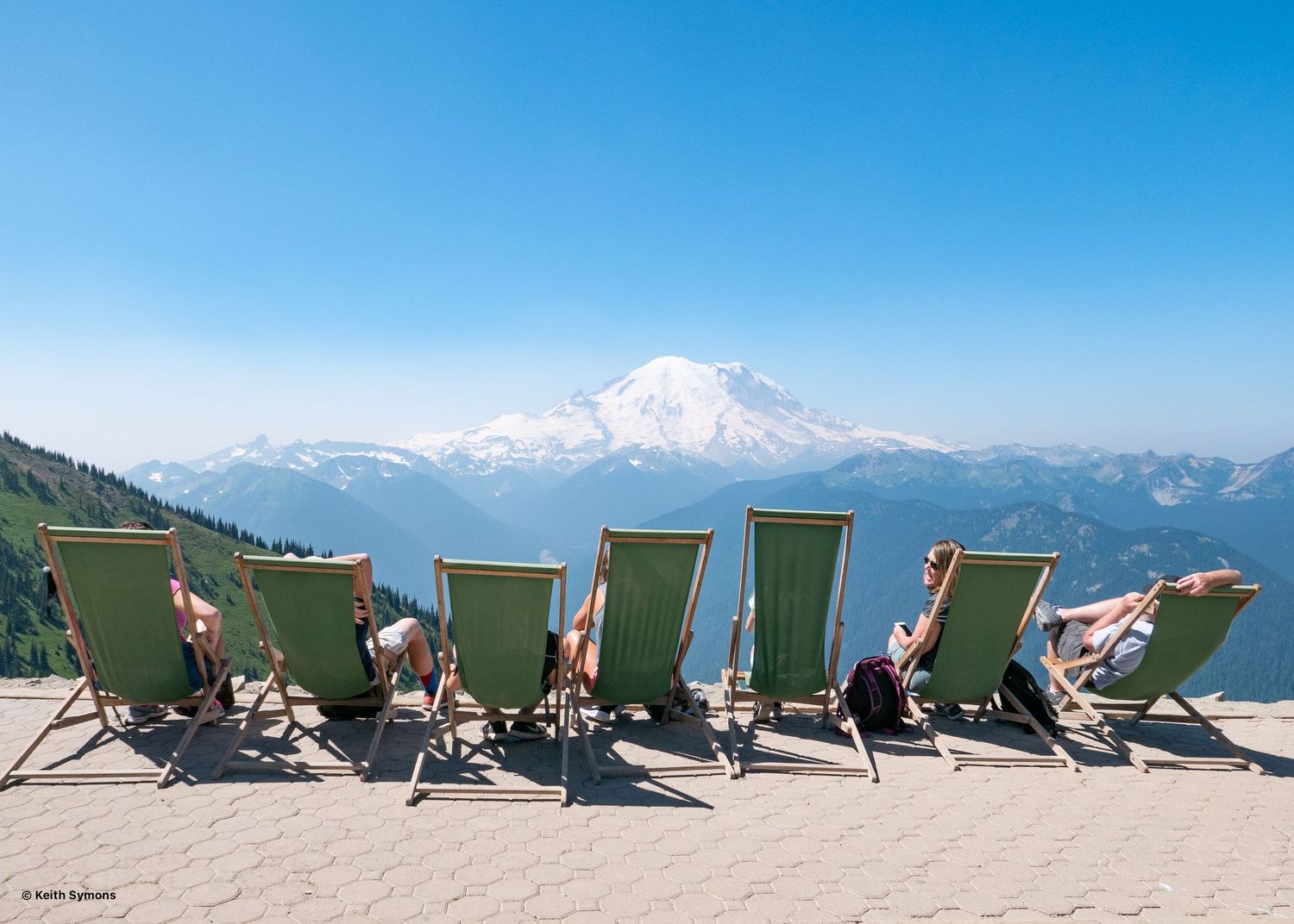 People relax in green lounge chairs on a mountain viewpoint overlooking snow-capped Mount Rainier on a clear day.
