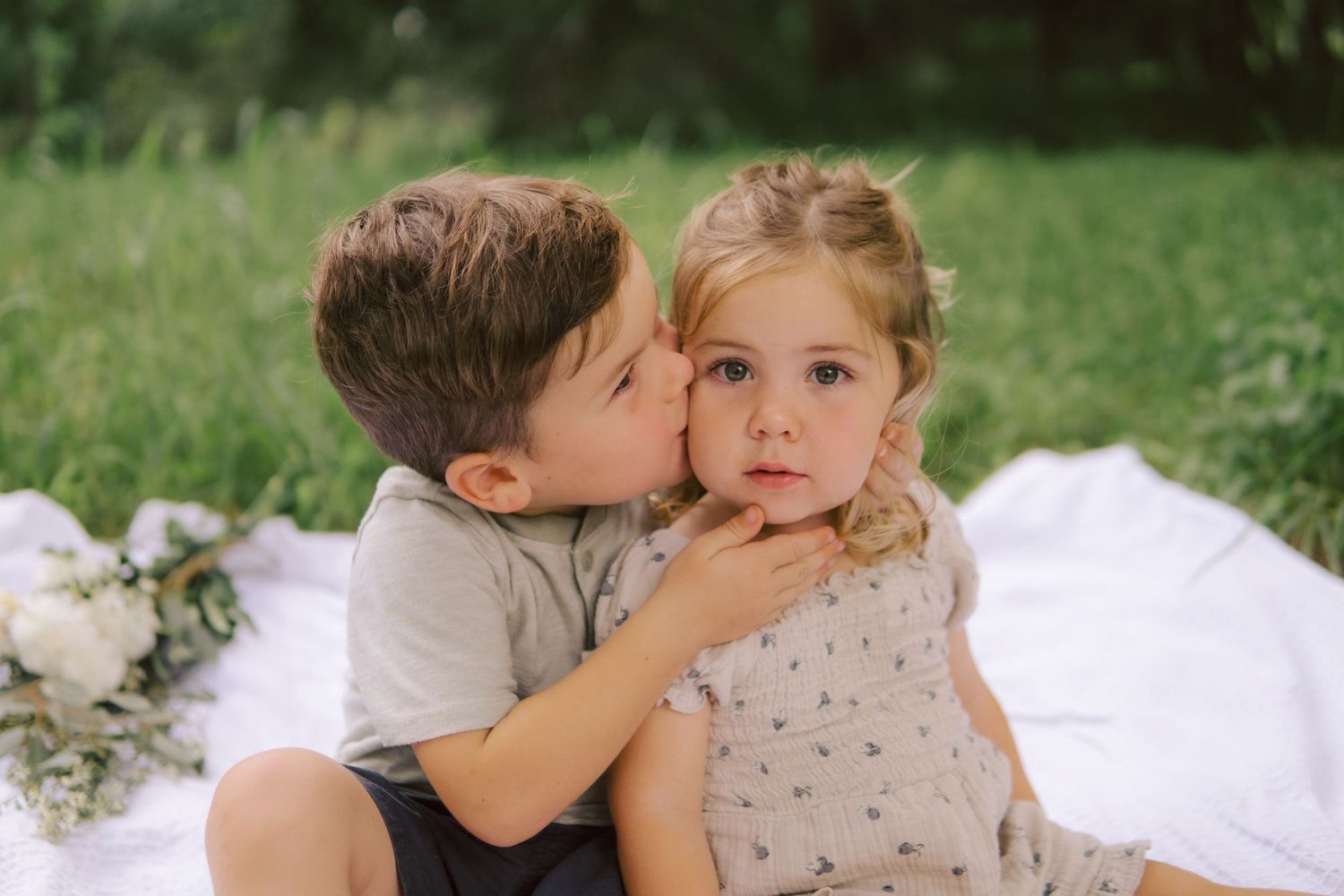 Two young children sitting together outdoors on a white blanket in a grassy field during summer in South Austin.