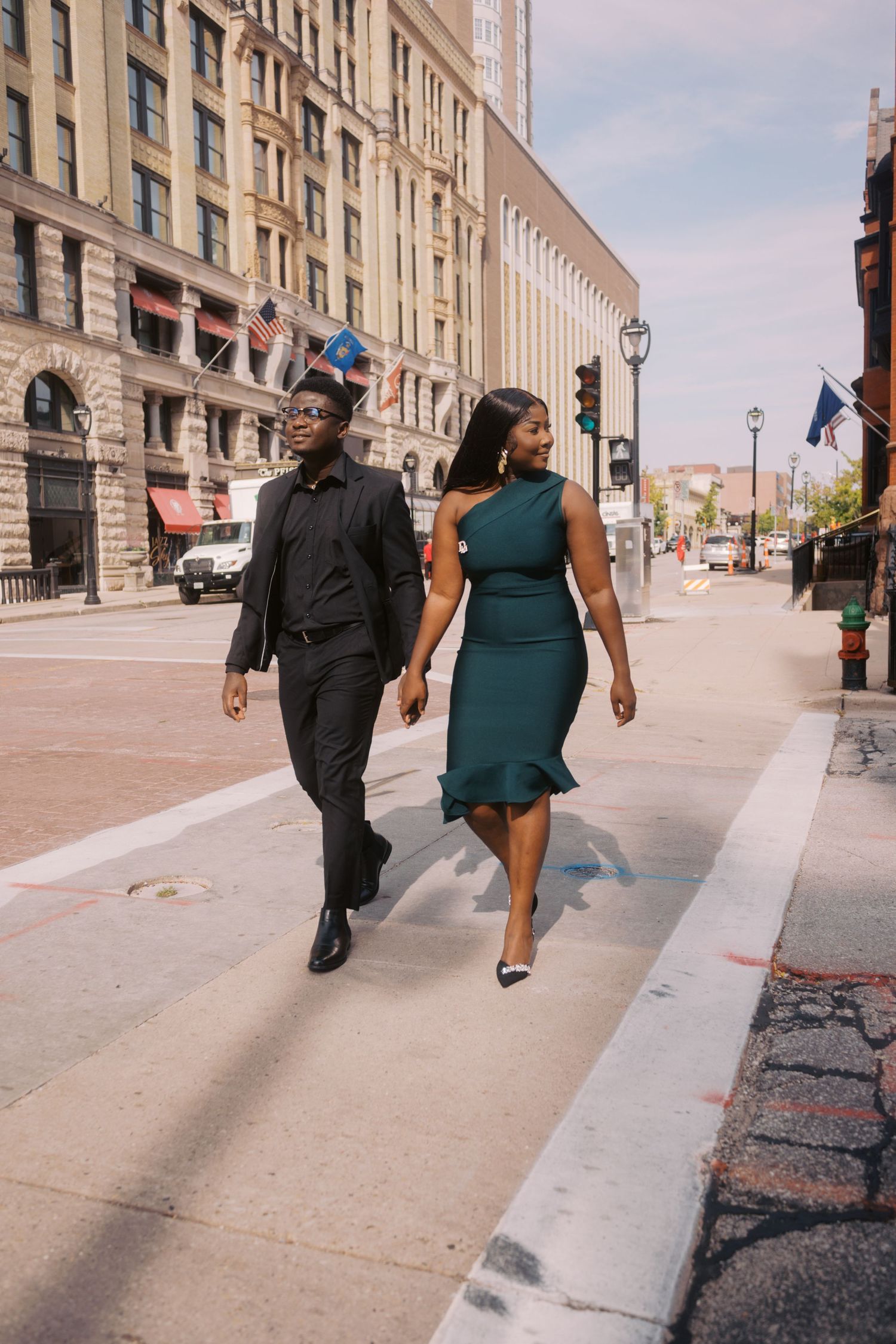 A couple walking together on a city street, wearing formal attire with classic architecture in the background.
