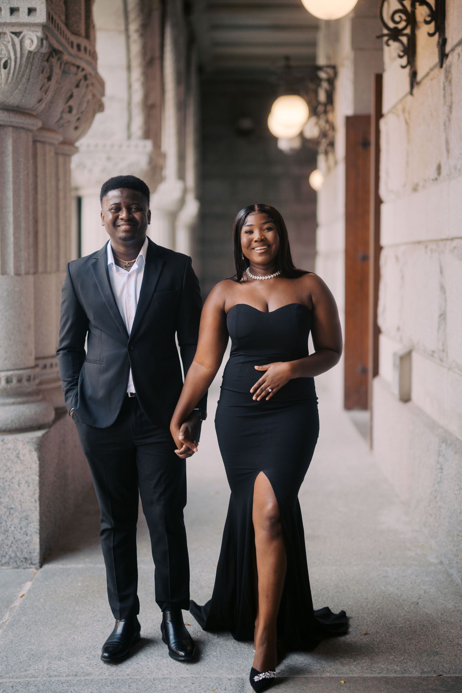 A couple in elegant formal attire poses together in a hallway with ornate architecture and glowing wall sconces.
