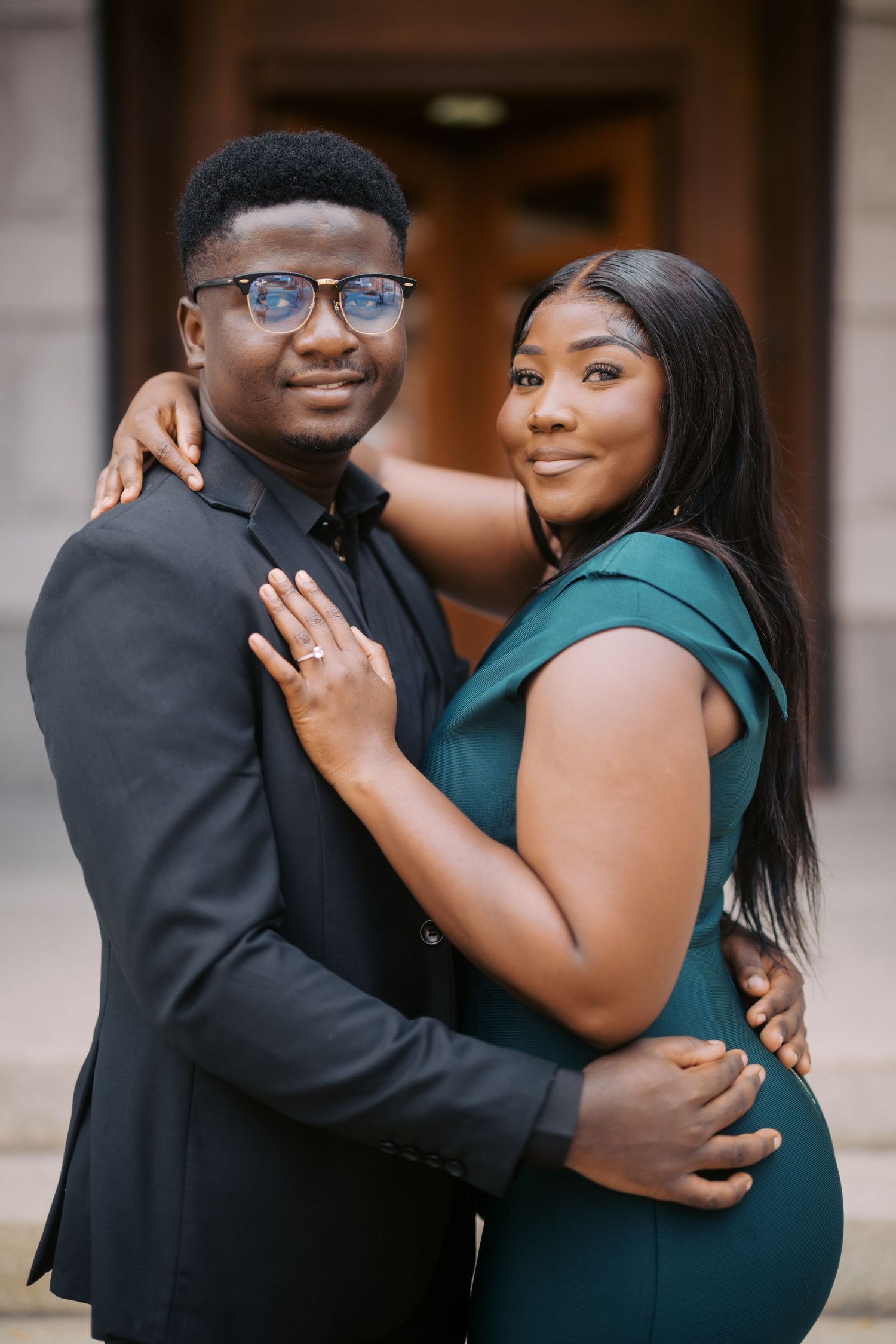 A loving couple embraces while wearing formal attire, with him in a black suit and her in an elegant green dress.
