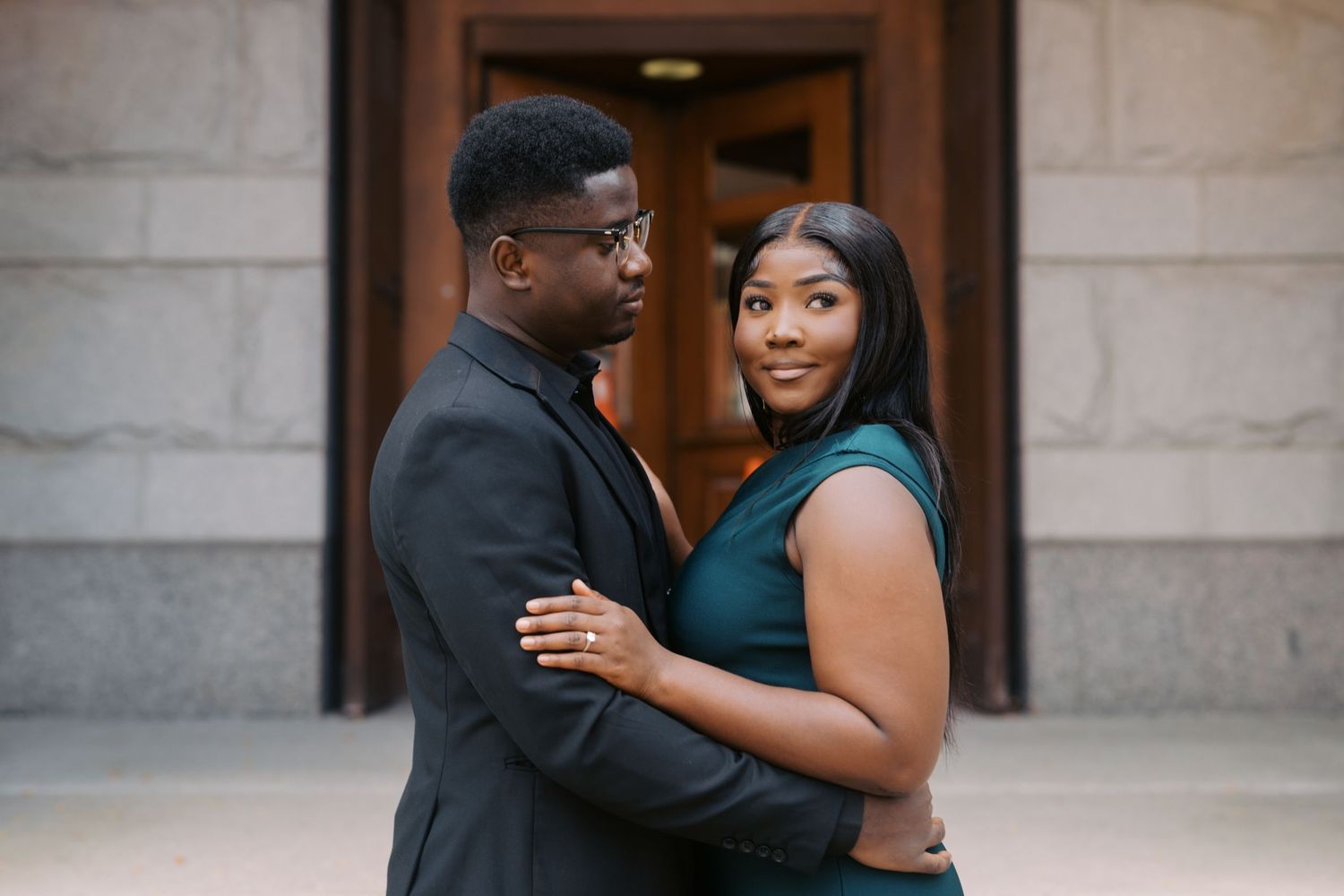 A couple stands close together smiling while posing outside a building with stone columns.