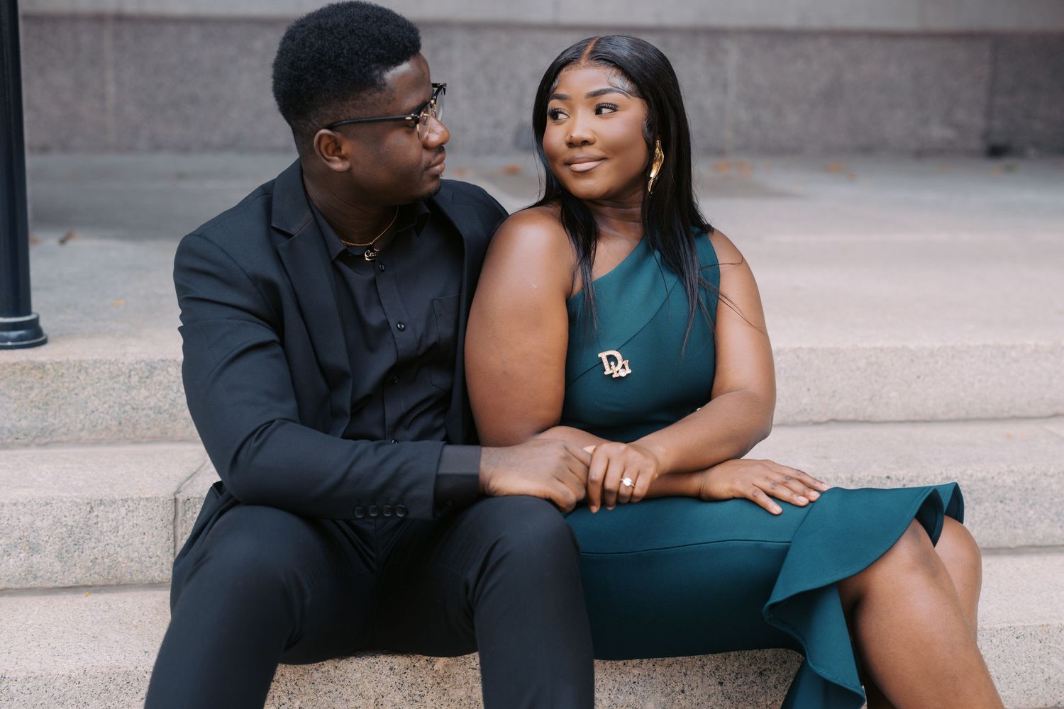 A couple sits together intimately on concrete steps, wearing formal attire in black and deep teal.