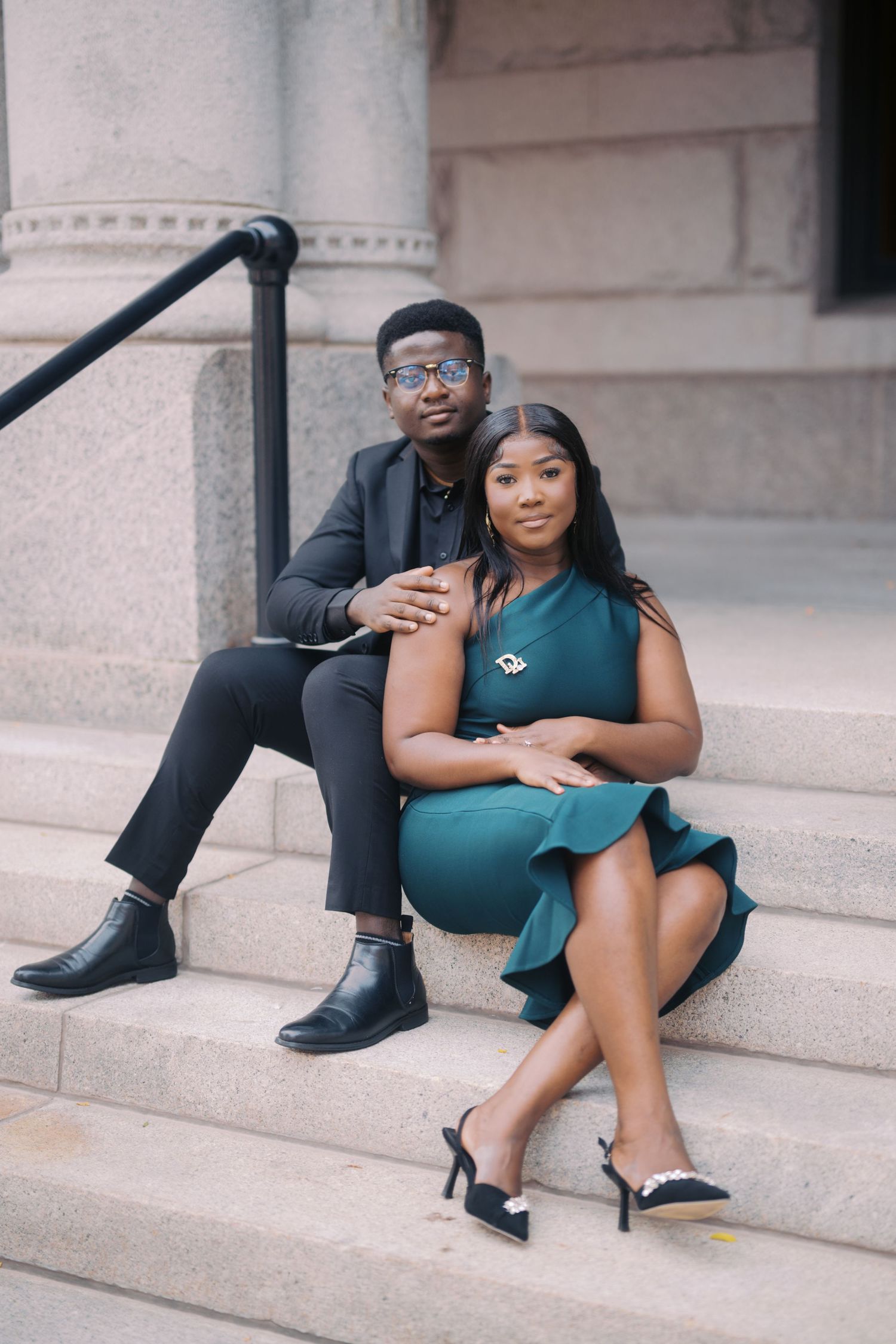 A couple sits together on outdoor stone steps, dressed elegantly with him in black and her in a teal dress.