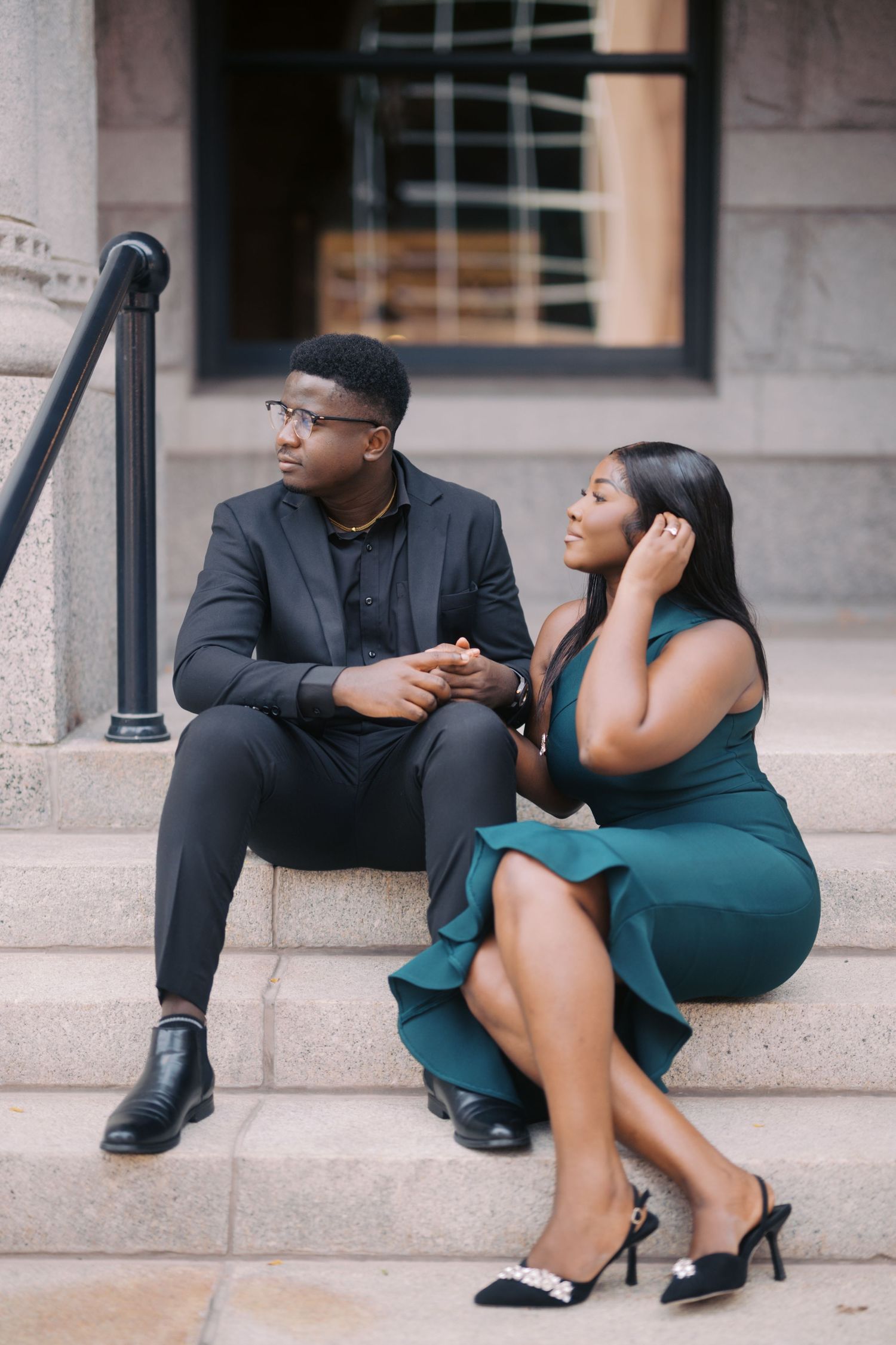 A stylishly dressed couple sits together on stone steps outside a brownstone building, wearing black and emerald outfits.
