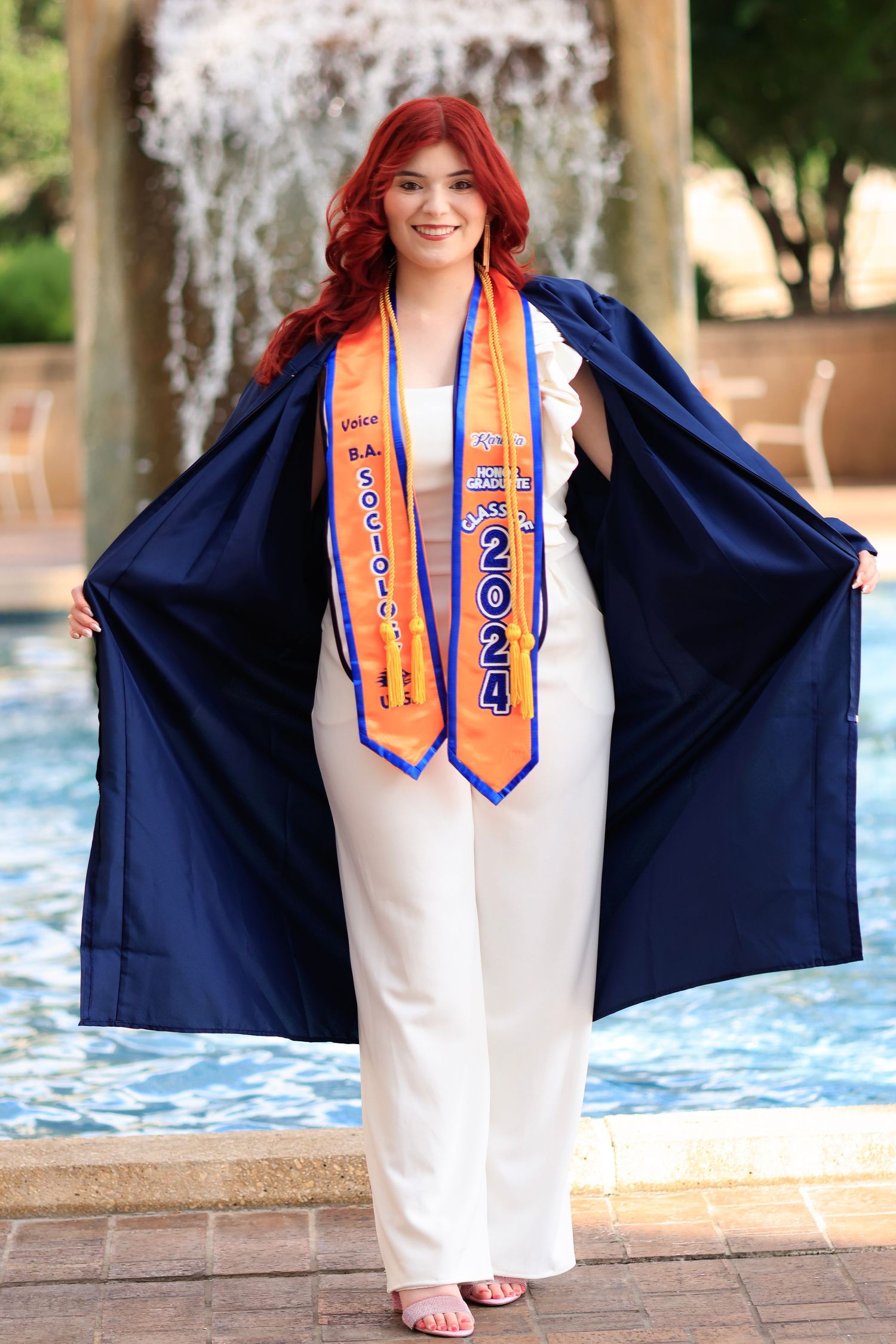Graduate in white outfit and navy cap displaying orange graduation stoles in front of fountain.