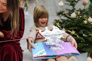 Person in red velvet dress reads interactive book with children in matching sweaters near Christmas tree.