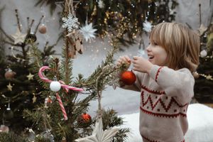Small child decorates Christmas tree with ornaments while wearing festive knit sweater.