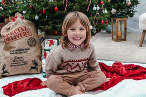 Child in holiday sweater poses by Christmas tree with special delivery sack nearby.