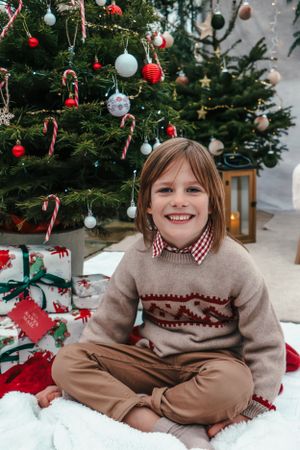Child in fair isle sweater sits cross-legged by Christmas tree surrounded by candy canes and ornaments.